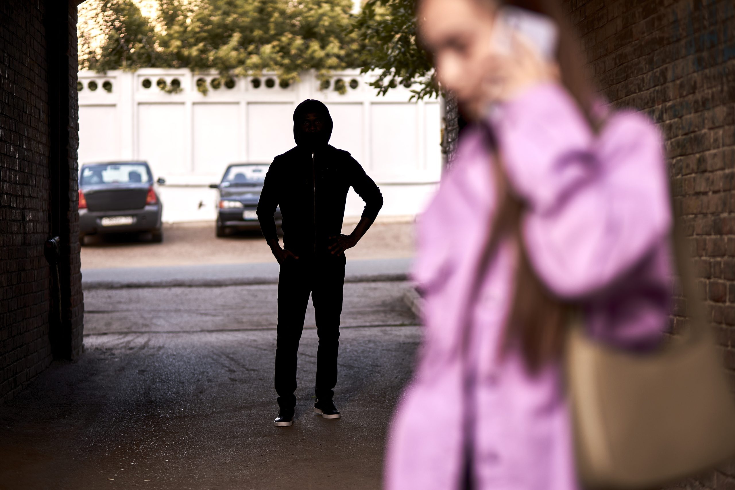 Jonge man met hoodie in donker tunneltje, op voorgrond vage afbeelding van een vrouw met roze jas die belt met haar mobiel. Ter illustratie van het CCV thema High Impact Crimes.