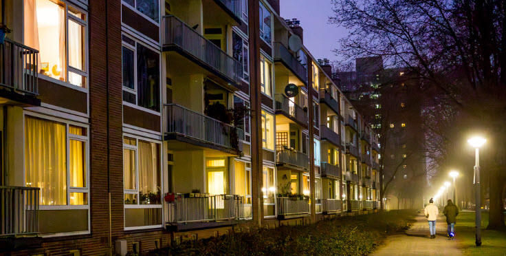 twee mensen lopen 's avonds over stoep langs flatgebouw.
