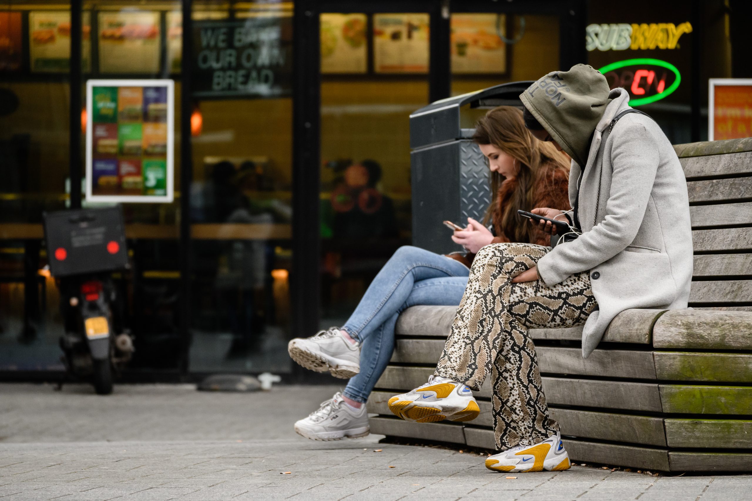 2 jongeren zitten op een bankje in een winkelcentrum op hun mobiel te kijken. Ter illustratie van dossiers over jongeren en veiligheid van het CCV.