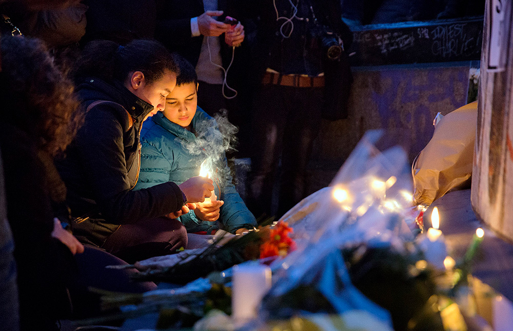 2 kinderen branden een kaarsje bij een bloemenzee om slachtoffers van een aanslag te herdenken.