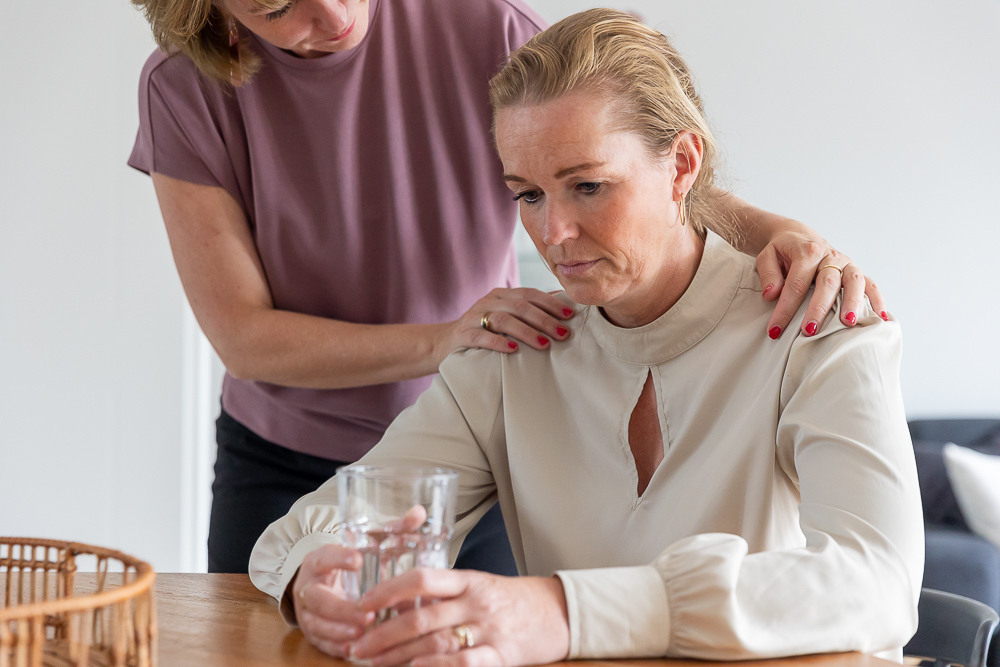 Vrouw legt haar handen op de schouder van een andere vrouw die verdrietig aan een tafel zit. Illustratie van het CCV bij het onderwerp Veiligheid en Zorg.